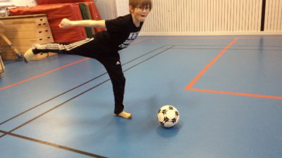 Noel står i en idrottshall med ett höjt ben och ska precis sparka på en fotboll.