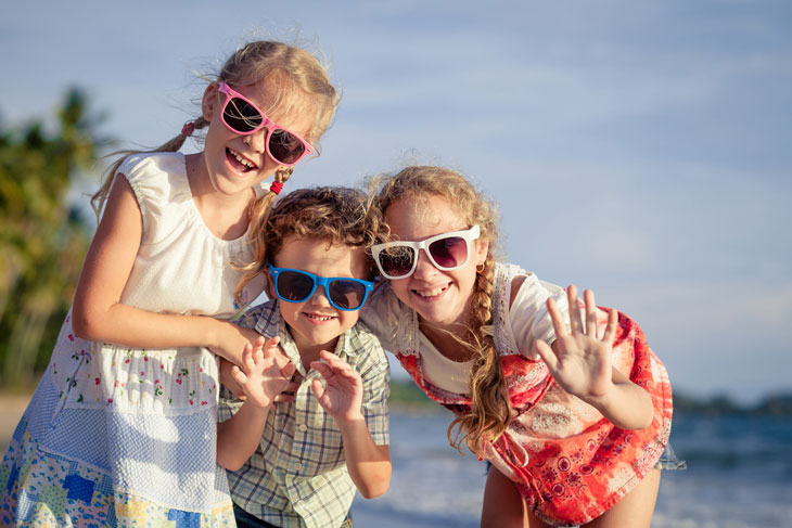 Tre syskon i solglasögon med olika färger ler framför havet.