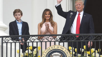 Donald Trum står på vita husets balkong och vingar med en hand. Bredvid honom står Melania som klappar händerna. Bredvid Trump och Melania står deras son.