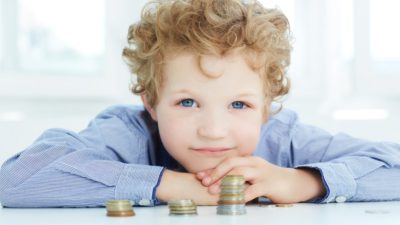 Ett barn med lockigt hår och blå ögon vilar sitt huvud på sina händer. Händerna ligger på ett bord och framför barnet på bordet ligger några högar med mynt.