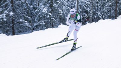 Charlotte åker skidor fort fram genom en snötäckt skog. Hon har färgglada skidor och vita tajta kläder. Hon har mössa, gula glasögon och håller stavarna under armarna så de pekar bakåt.