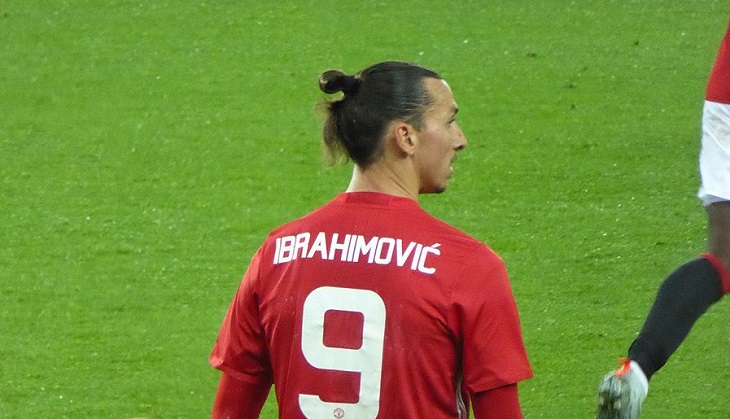 Zlatan står på en fotbollsplan med ryggen mot oss. Han tittar åt sidan och har en röd fotbollströja på sig. På ryggen står det Ibrahimovic och siffran 9 i vitt.