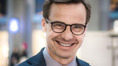 Ulf Kristersson har glasögon, kort hår och en kostym. Han har en del rynkor runt ögonen som syns extra mycket eftersom han ler stort.
