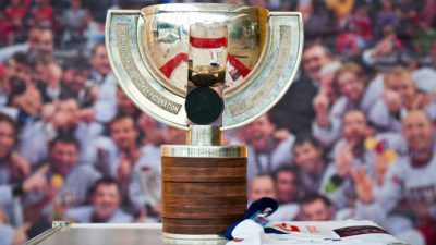 En stor guldig pokal står på ett bort. Pokalen har en fot av trä, en stor skål av guld. Från foten och upp runt var sin sida av skålen går två remsor av metal ut. På remsorna står det text.