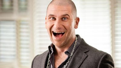 Markoolio ler med en jättestor öppen mun. Han har kavaj och skjorta på sig, och en stor kedja runt halsen. Han har snaggat hår och glada ögon. Han ser lite galen ut.