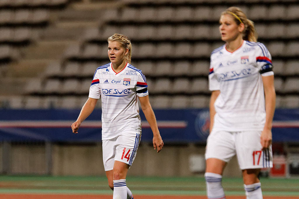 Ada går över en fotbollsplan. Hon har fotbollskläder på sig och sitt hår uppsatt i en tofs. I bakgrunden syns fotbollsläktare.