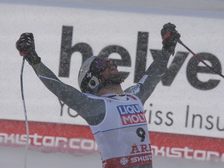 Aksel har skidklädsel, hjälm och skidglasögon på sig. I händerna håller han sina stavar som han sträcker upp i luften som en segergest.