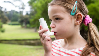 Ett barn står utomhus. Det har långt hår med tofsar och spännen i. Barnet håller en avlång plastgrej framför munnen